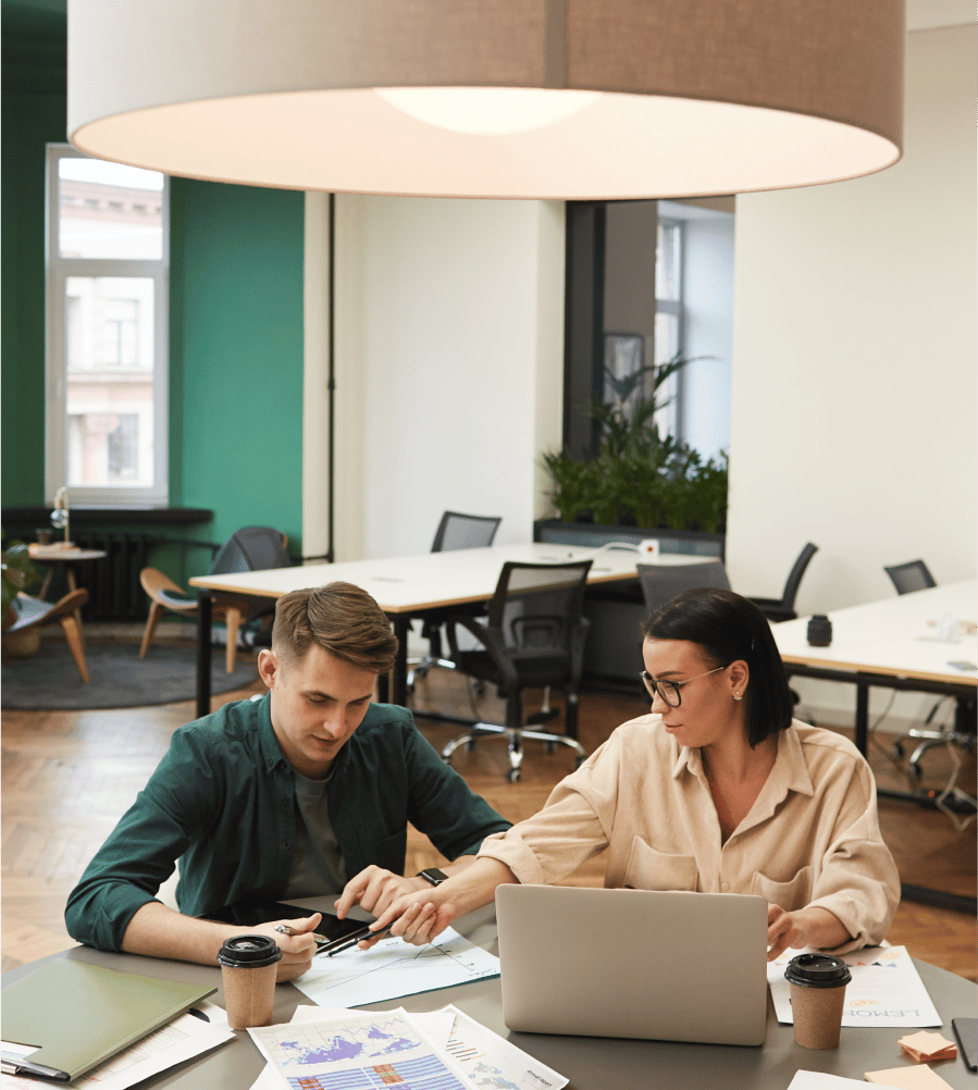 A man and woman sit in an open concept office with coffee and work in front of them