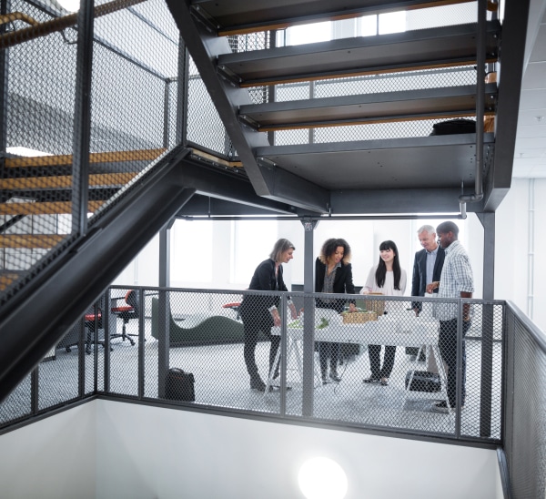 A team of 5 meet around a large white able behind an industrial staircase in the office