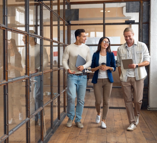 3 colleagues walk down a hall holding a laptop and coffee and smiling