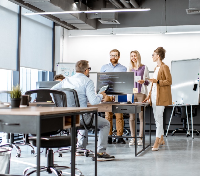 Team stands around a computer while colleague discusses work with them